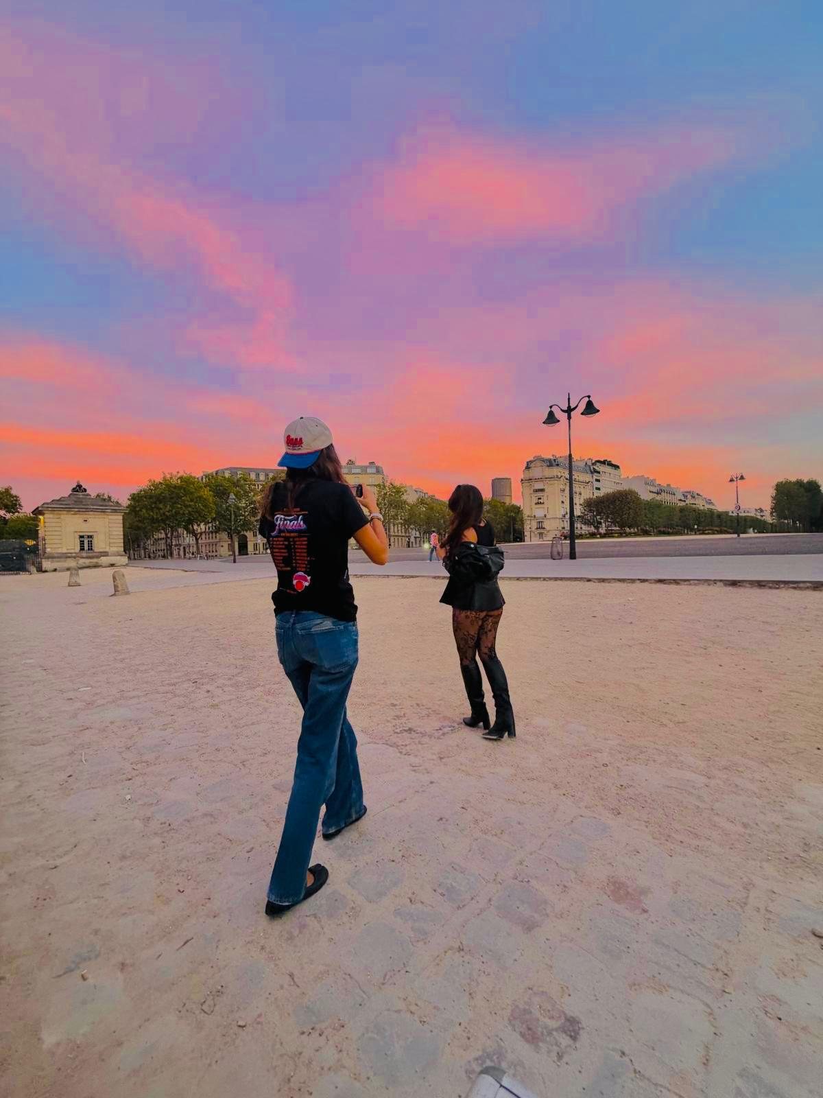 SEYA et une amie de dos sur une esplanade parisienne, sous un ciel rose et orangé au coucher du soleil